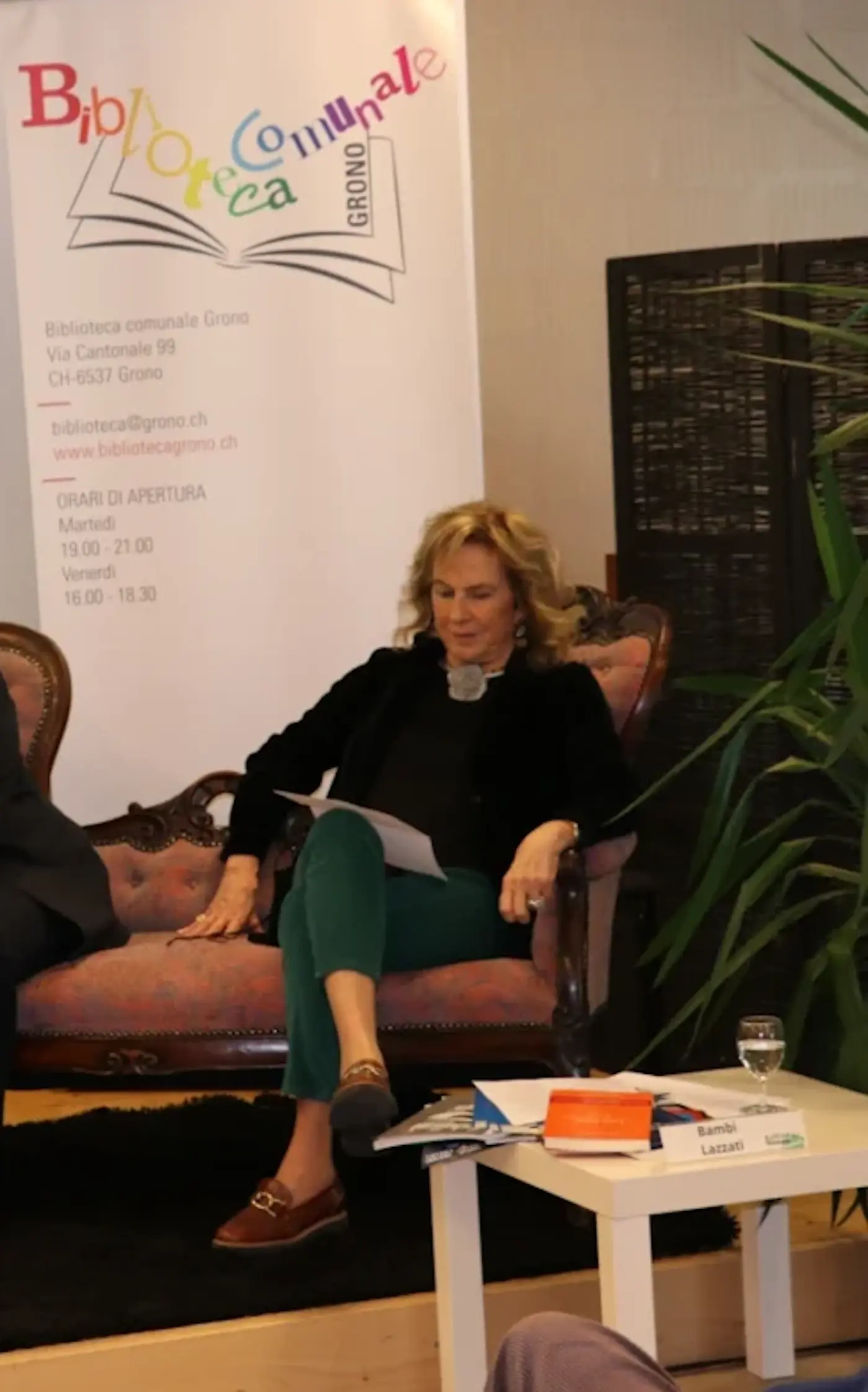 Una donna seduta su un divano elegante mentre legge appunti, circondata da piante verdi e un ambiente accogliente. Sullo sfondo, un banner con il logo della Biblioteca