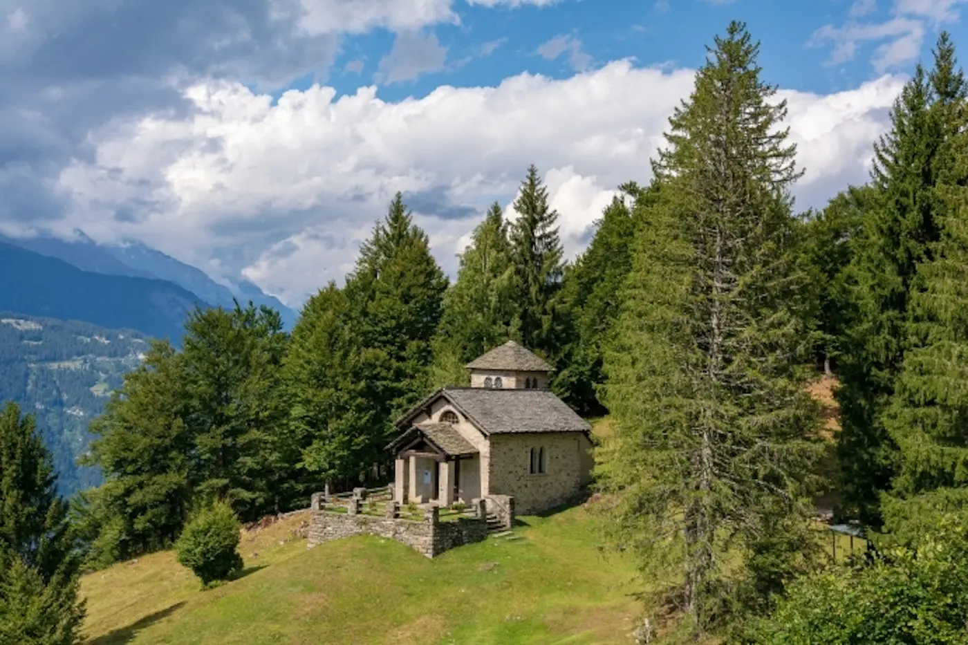 Una piccola chiesa in pietra situata su una collina verdeggiante, circondata da alberi alti e uno splendido cielo blu con nuvole bianche.