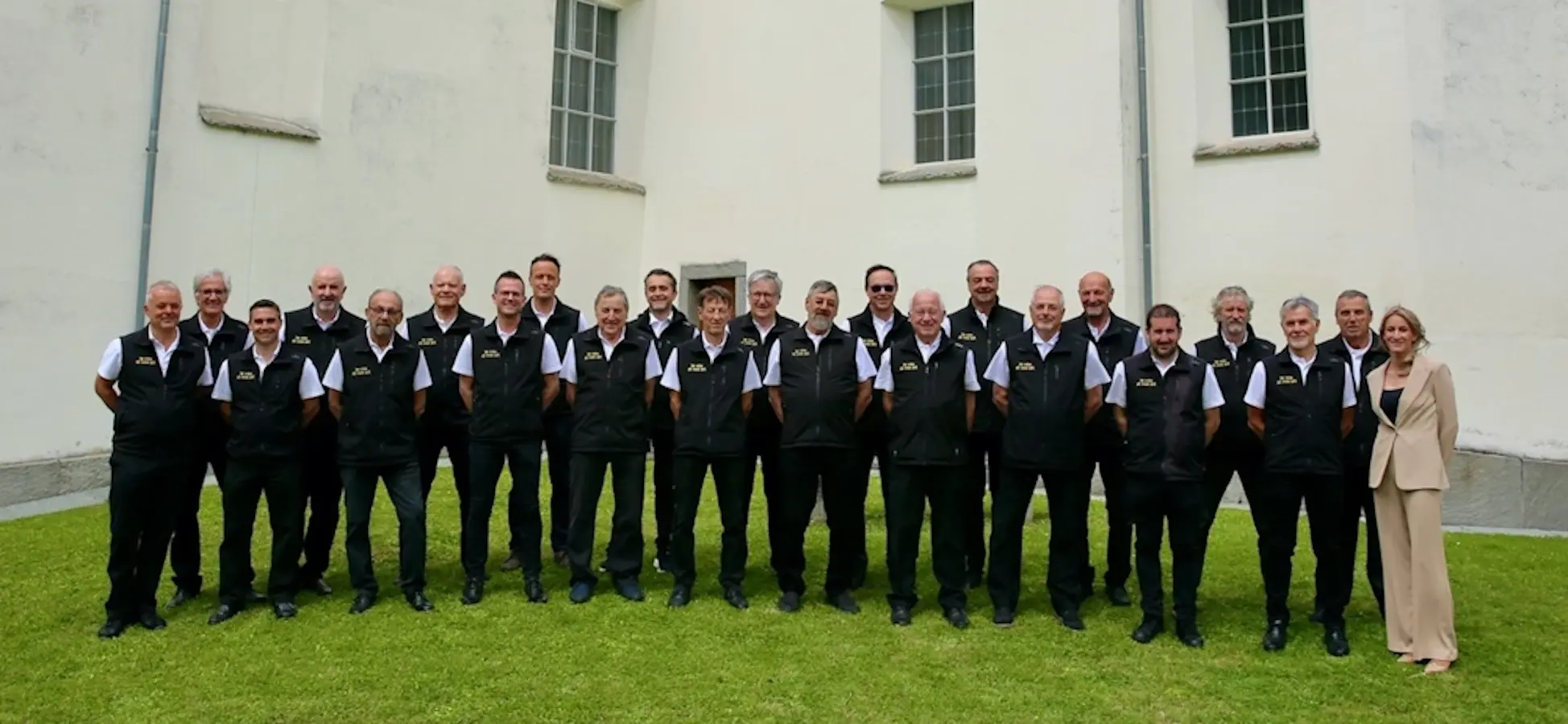 Gruppo di uomini in abbigliamento uniforme, schierati in fila su un prato verde, con un edificio bianco sullo sfondo.