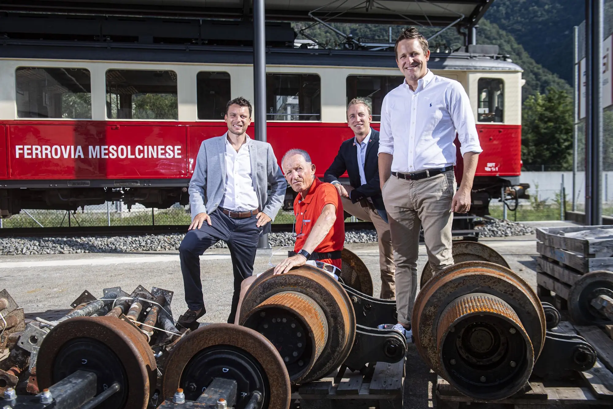 Quattro uomini sorridenti posano davanti a un treno rosso in un ambiente industriale. Sono circondati da rotaie e pezzi di treno. Un uomo anziano &egrave; sed