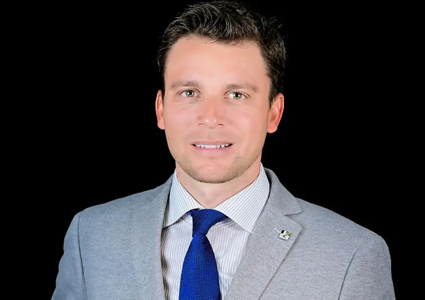 Uomo sorridente con giacca grigia e cravatta blu, su sfondo nero.