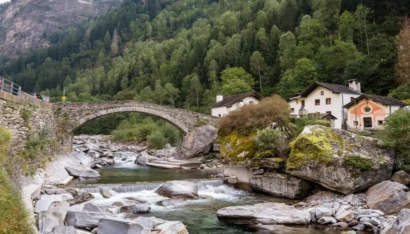 Un ponte in pietra attraversa un fiume circondato da rocce e vegetazione lussureggiante. Sullo sfondo si vedono case in stile tradizionale mont
