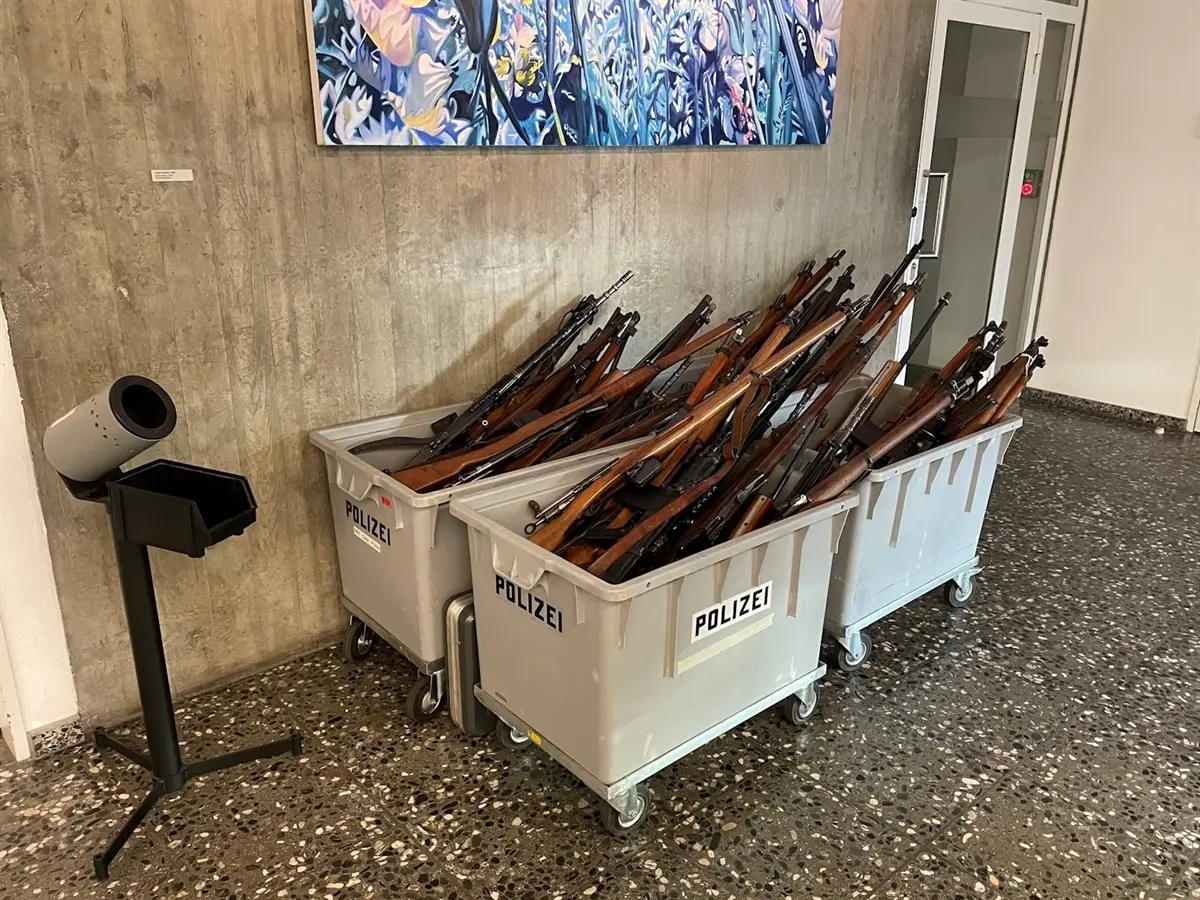 Fucili impilati all'interno di contenitori grigi con la scritta "POLIZEI" in un ambiente con pareti di cemento e un'opera d'arte appesa.