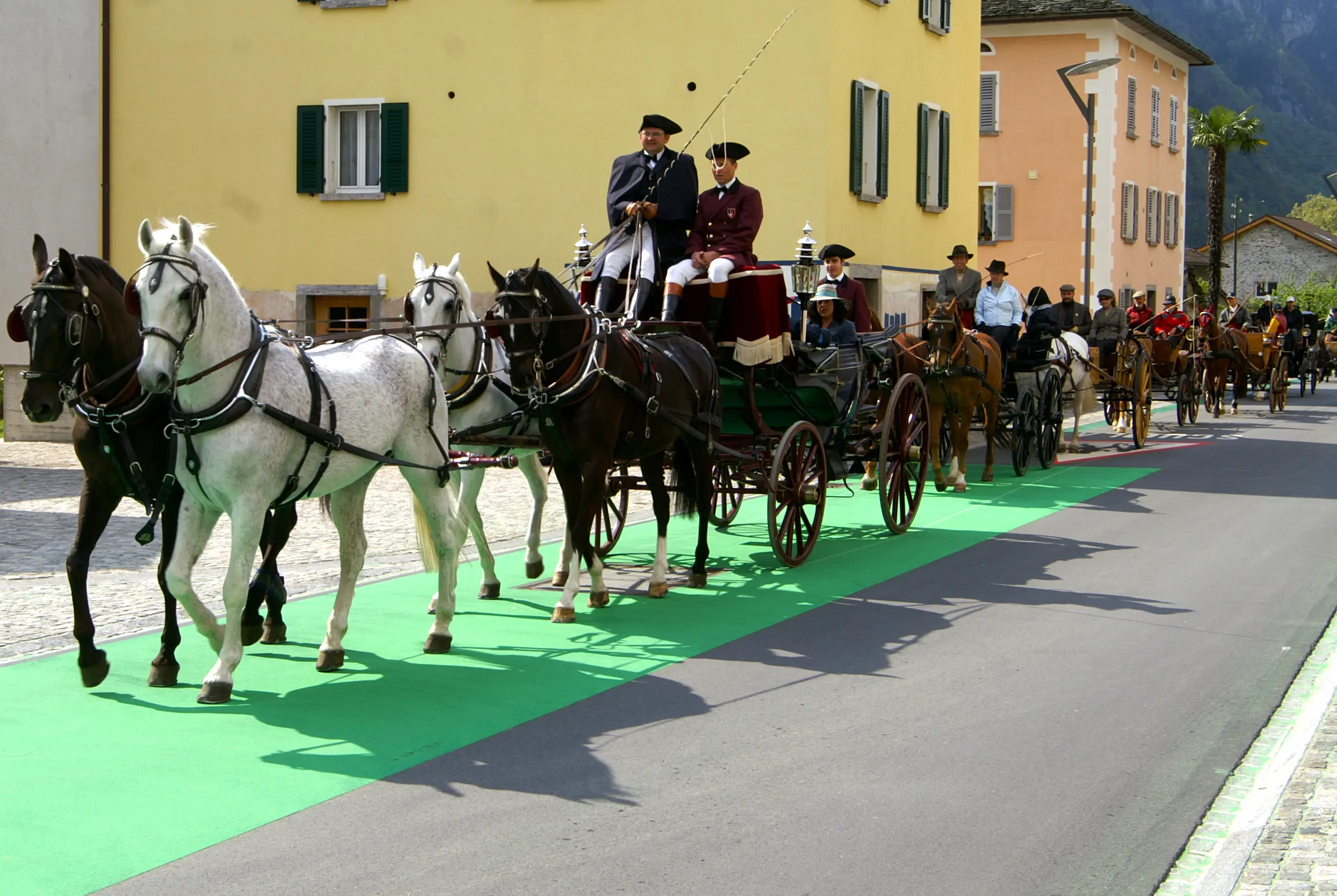 Una parata di carrozze trainate da cavalli su un tappeto verde, con persone in abbigliamento tradizionale. Sullo sfondo si vedono edifici storici e una montagna.