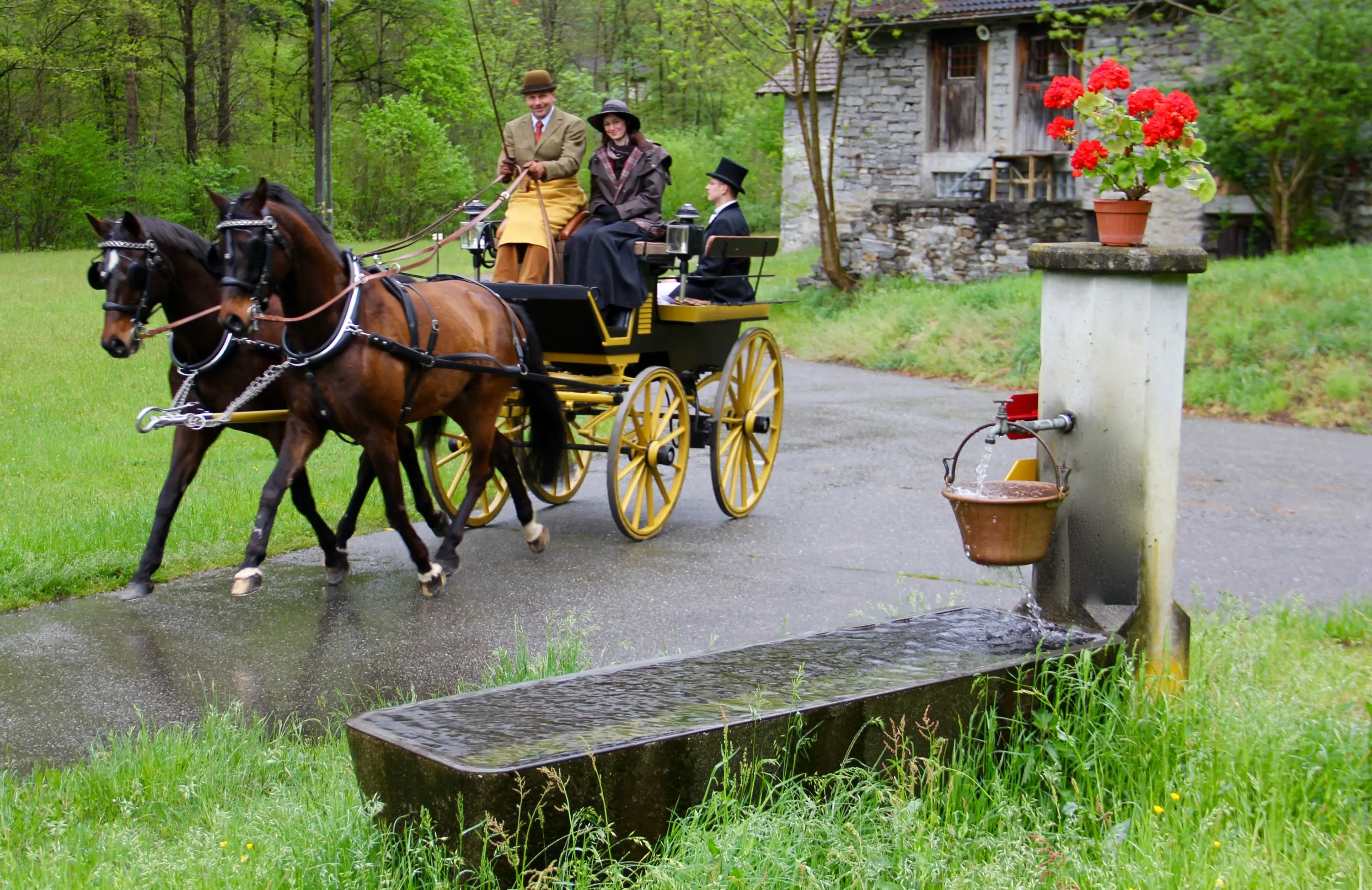 Una carrozza trainata da due cavalli attraversa un sentiero in un'area verde, con persone in abiti storici a bordo. In primo piano, un'antica fontana con un secchio appeso. Sullo sfondo, una casa in pietra circondata da alberi.