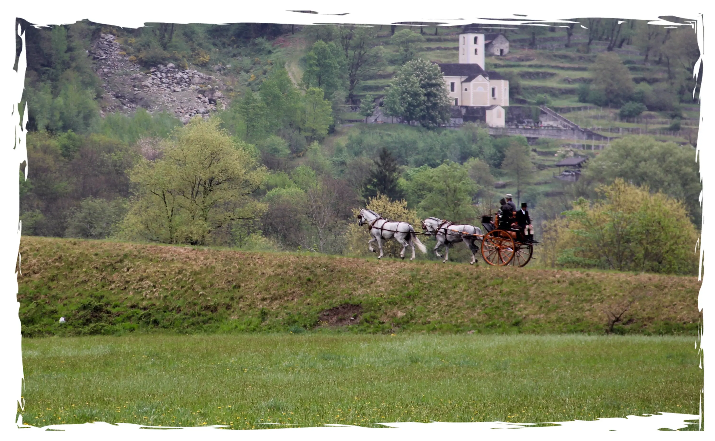Una carrozza trainata da cavalli passa attraverso un paesaggio verde, con una chiesa bianca visibile sullo sfondo tra gli alberi.