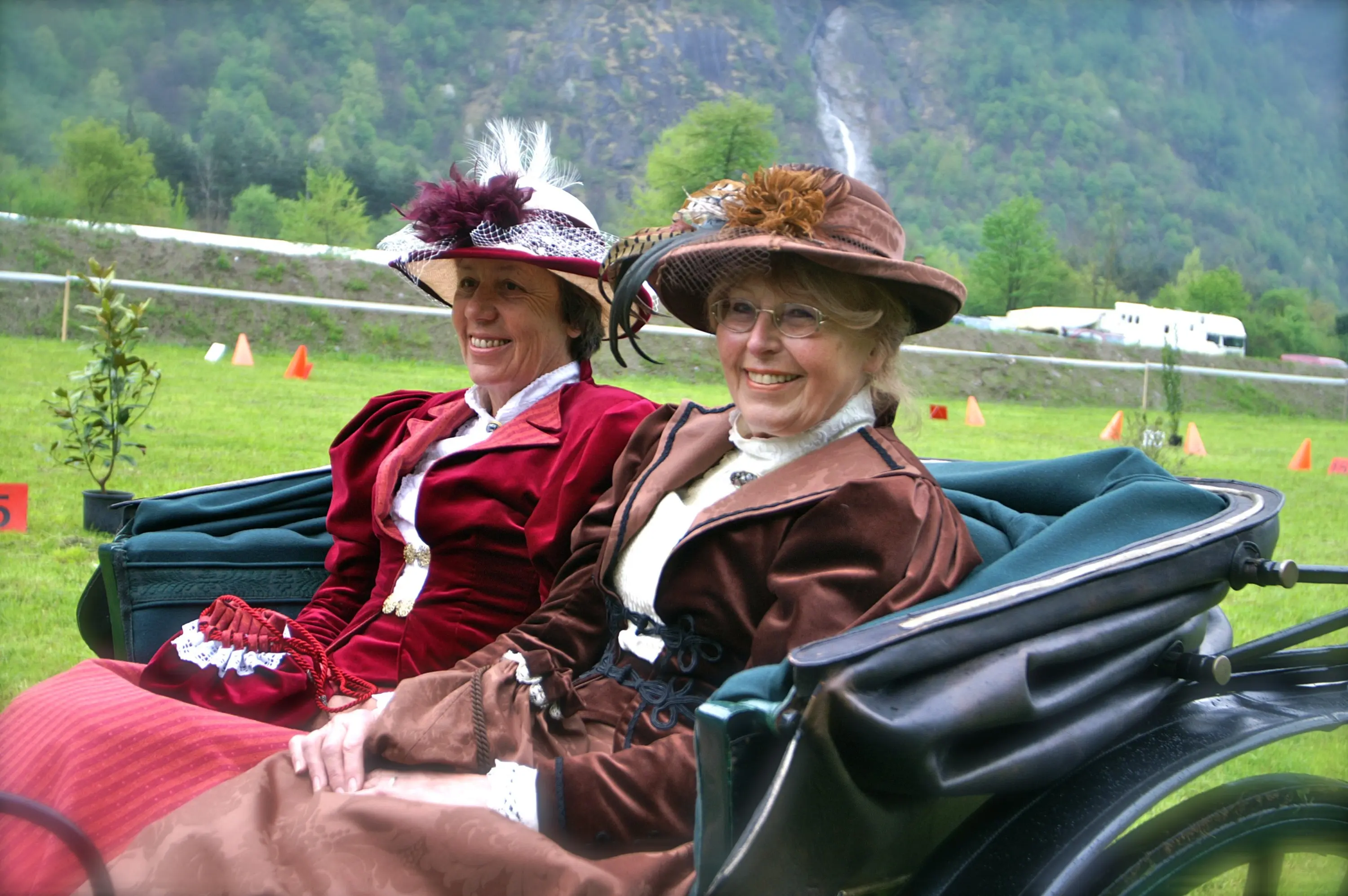 Due donne sorridenti, vestite in modo elegante con abiti vittoriani, si siedono su una carrozza all'aperto, circondate da una verdeggiante scena naturale.