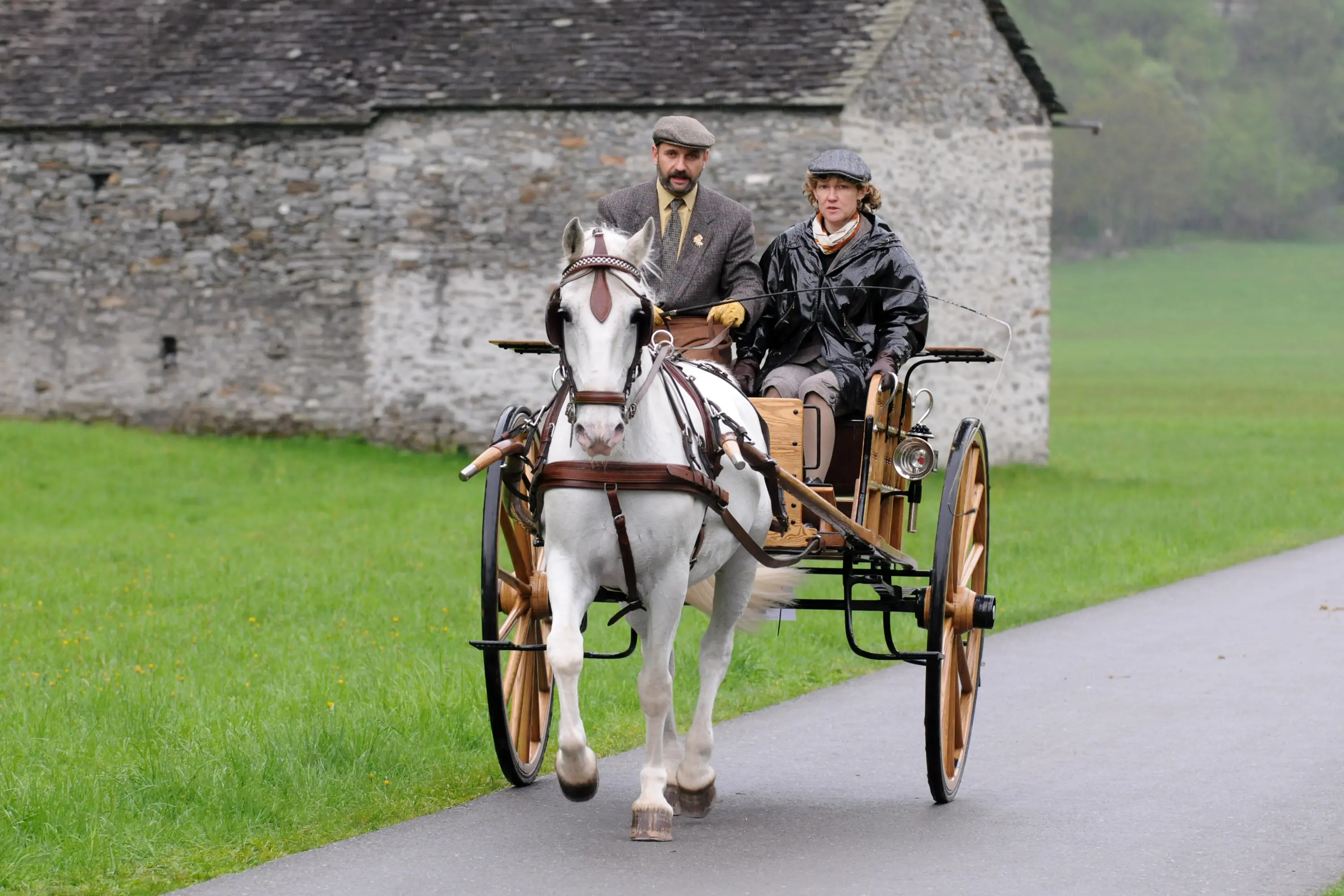 Due persone su un calesse trainato da un cavallo. L'uomo indossa un cappello e un abito elegante, mentre la donna è vestita con un impermeabile e pantaloni. Sullo sfondo si vede una vecchia costruzione in pietra e un prato verde.