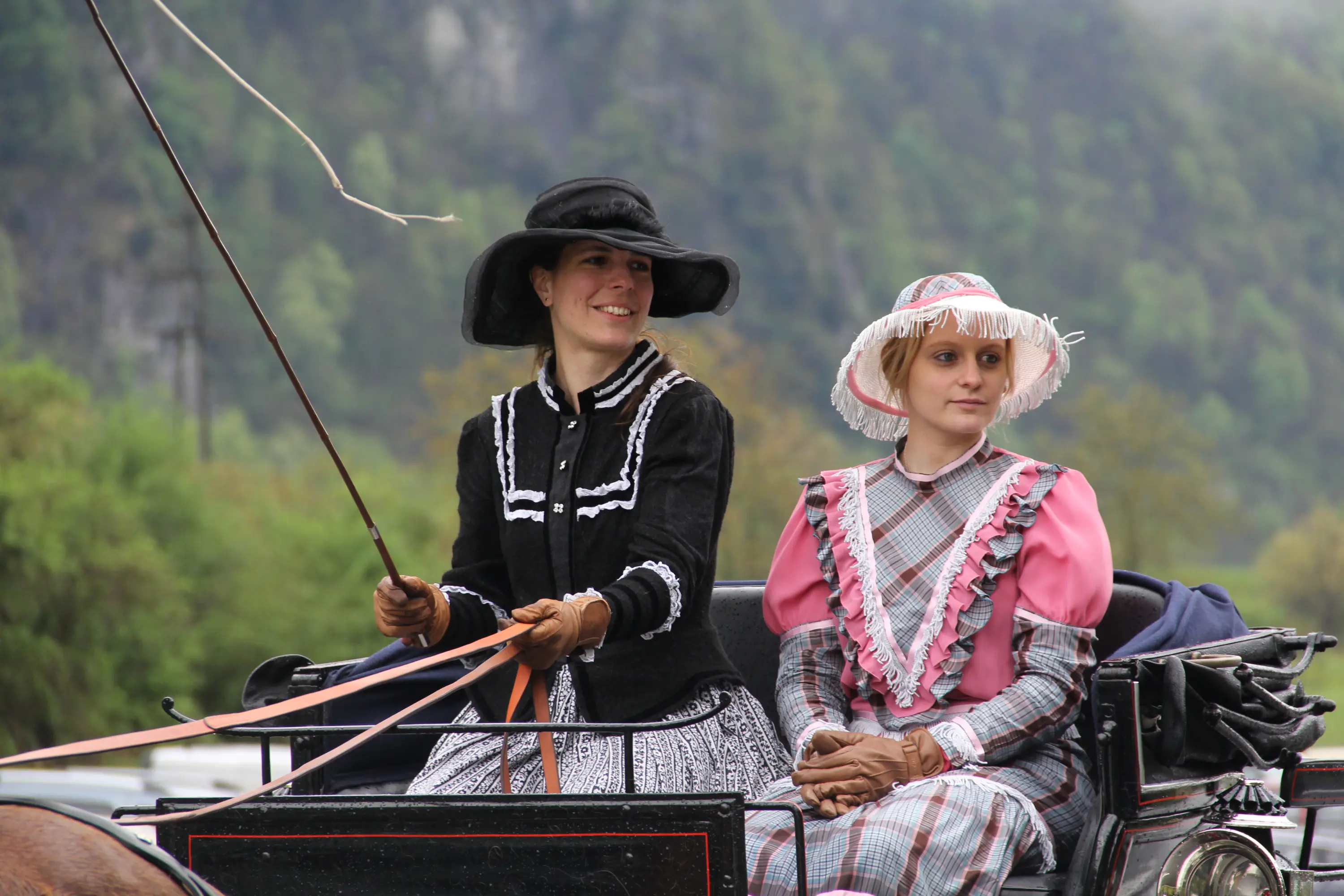 Due donne vestite in abiti storici sono sedute su un carrotrainato da cavalli. Una di loro tiene una frusta e indossa un cappello nero, mentre l'altra ha un cappello bianco e un abito rosa. Sullo sfondo si vedono alberi e colline verdeggianti.