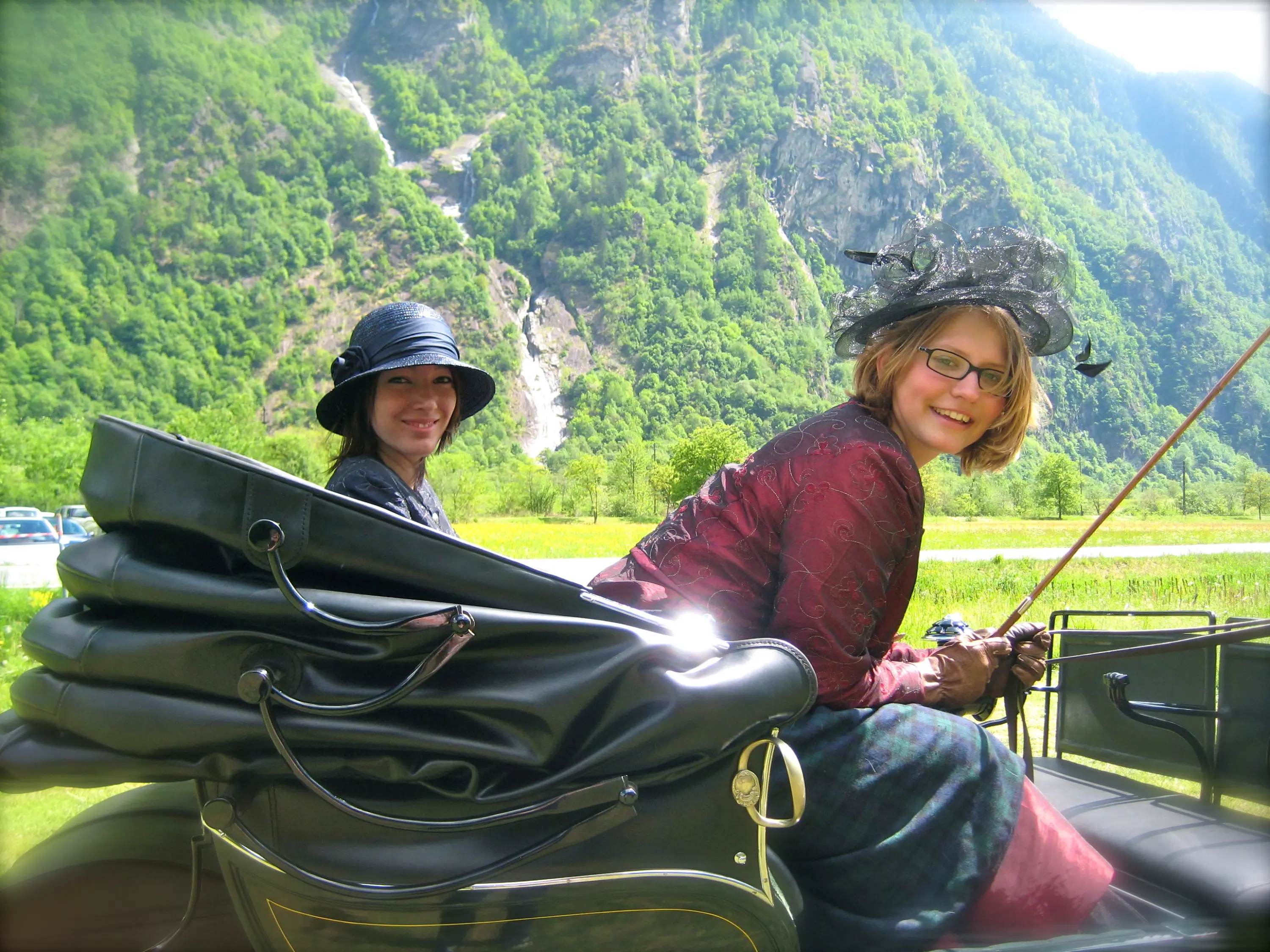 Due donne sorridenti sedute in una carrozza, circondate da una lussureggiante natura montana. Indossano abiti eleganti, uno dei quali ha un cappello a falda larga. Sullo sfondo si vedono alberi verdi e una cascata.