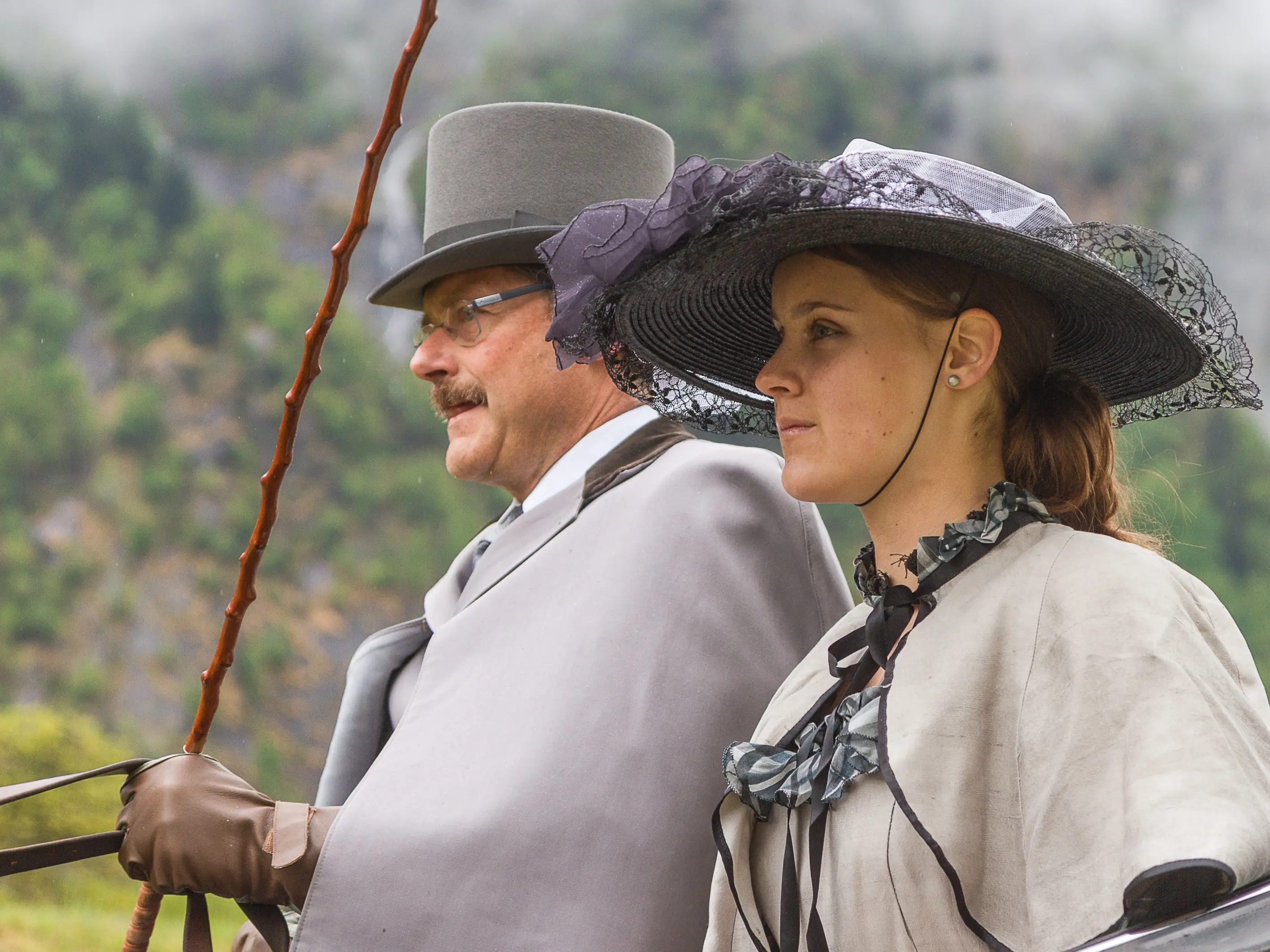 Due persone in abiti storici stanno in posa all'aperto. L'uomo indossa un cappello a cilindro e una giacca grigia, mentre la donna ha un cappello decorato con pizzo e un mantello chiaro. Sullo sfondo si intravede la natura.