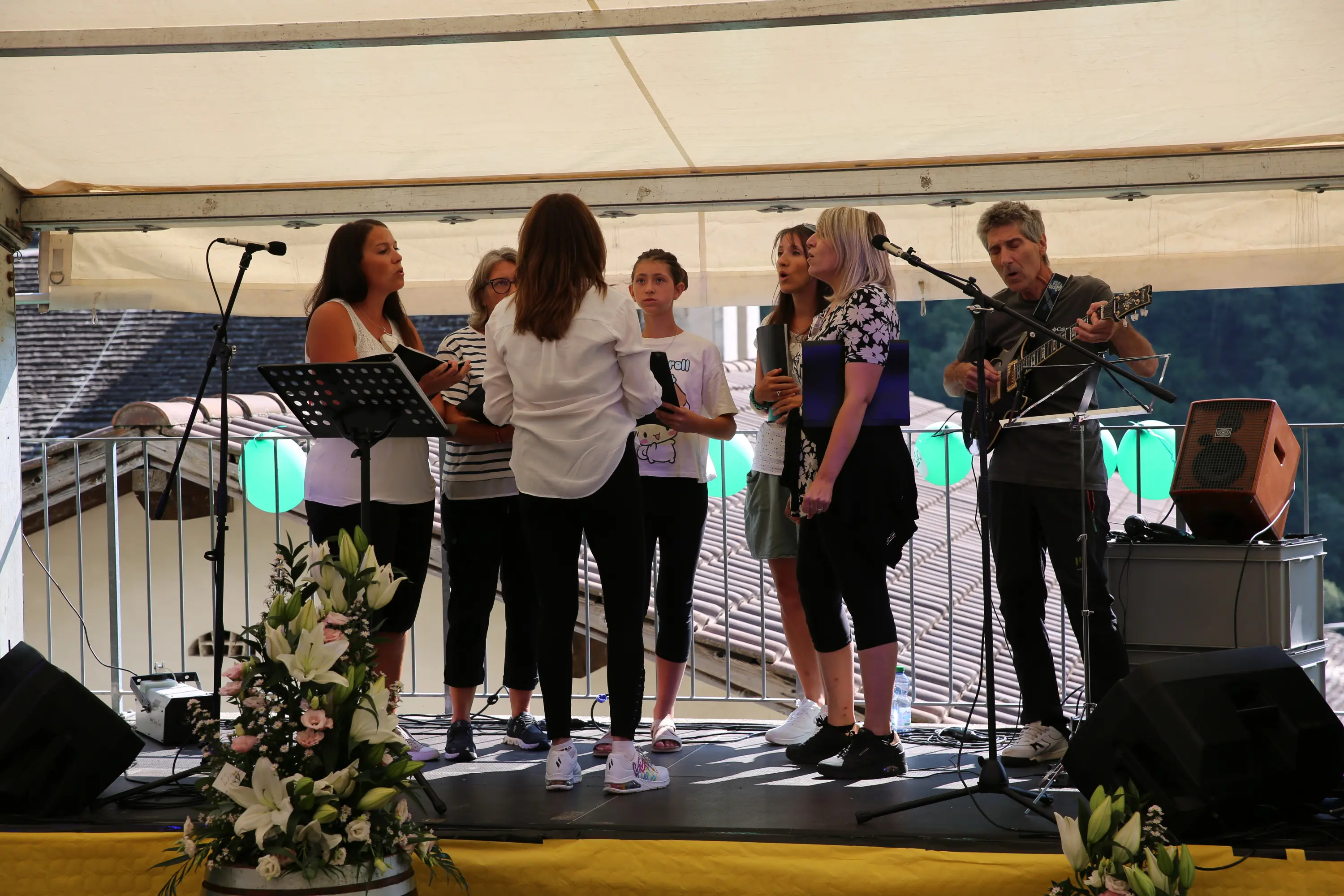 Un gruppo di sei persone si esibisce su un palco. Una cantante di spalle guida il gruppo, mentre gli altri cantano. Un musicista suona la chitarra. Sullo sfondo, un tendone bianco e decorazioni verdi. Fiori bianchi e un microfono adornano il palco.