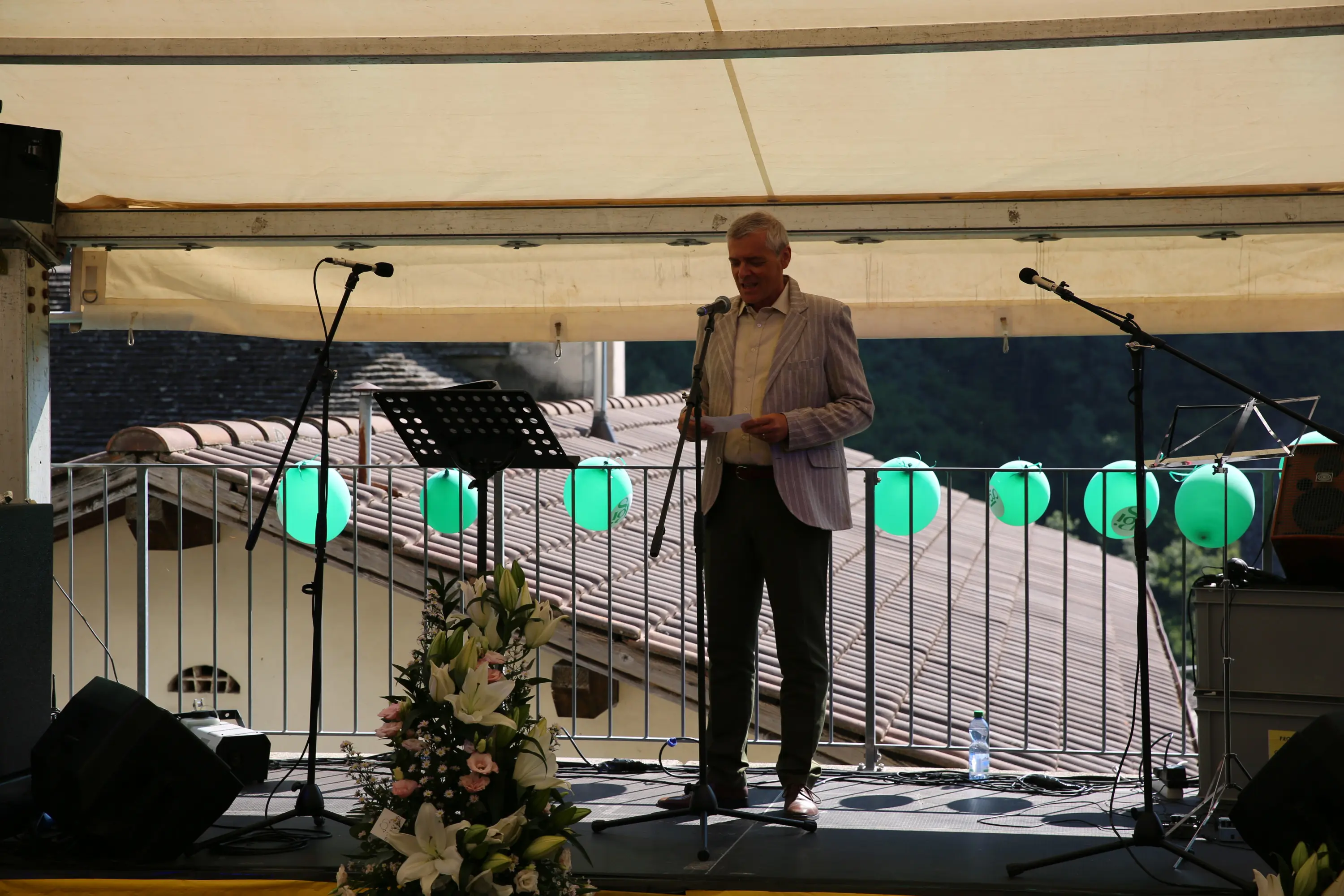 Un uomo in giacca a righe parla al microfono su un palco all'aperto, con decorazioni di palloncini verdi e un bouquet di fiori bianchi e rosa accanto a lui.