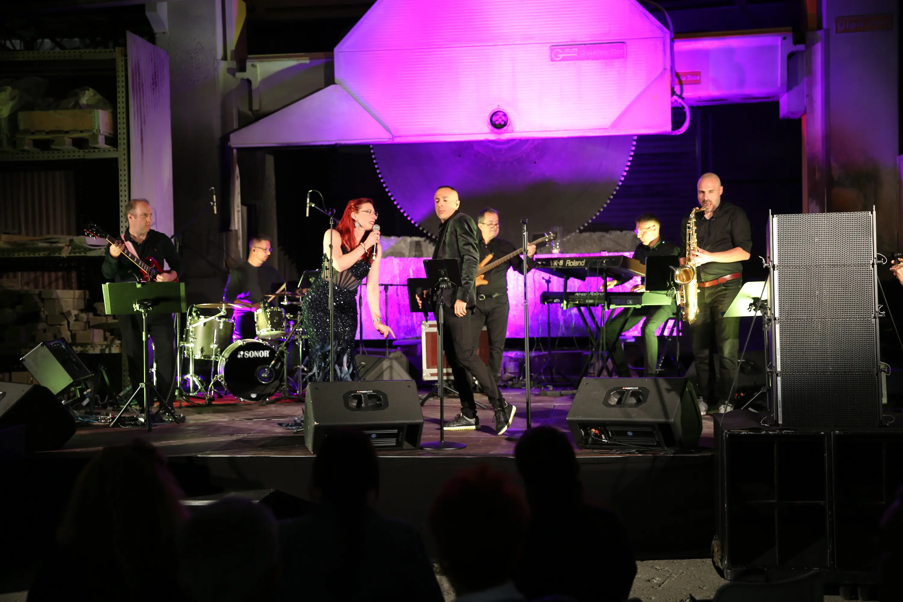 Una band si esibisce su un palco illuminato da luci violette. I musicisti includono cantanti, un sassofonista, e diverse strumentazioni come batteria e tastiere. Sullo sfondo si trova un grande oggetto che assomiglia a una sega industriale.