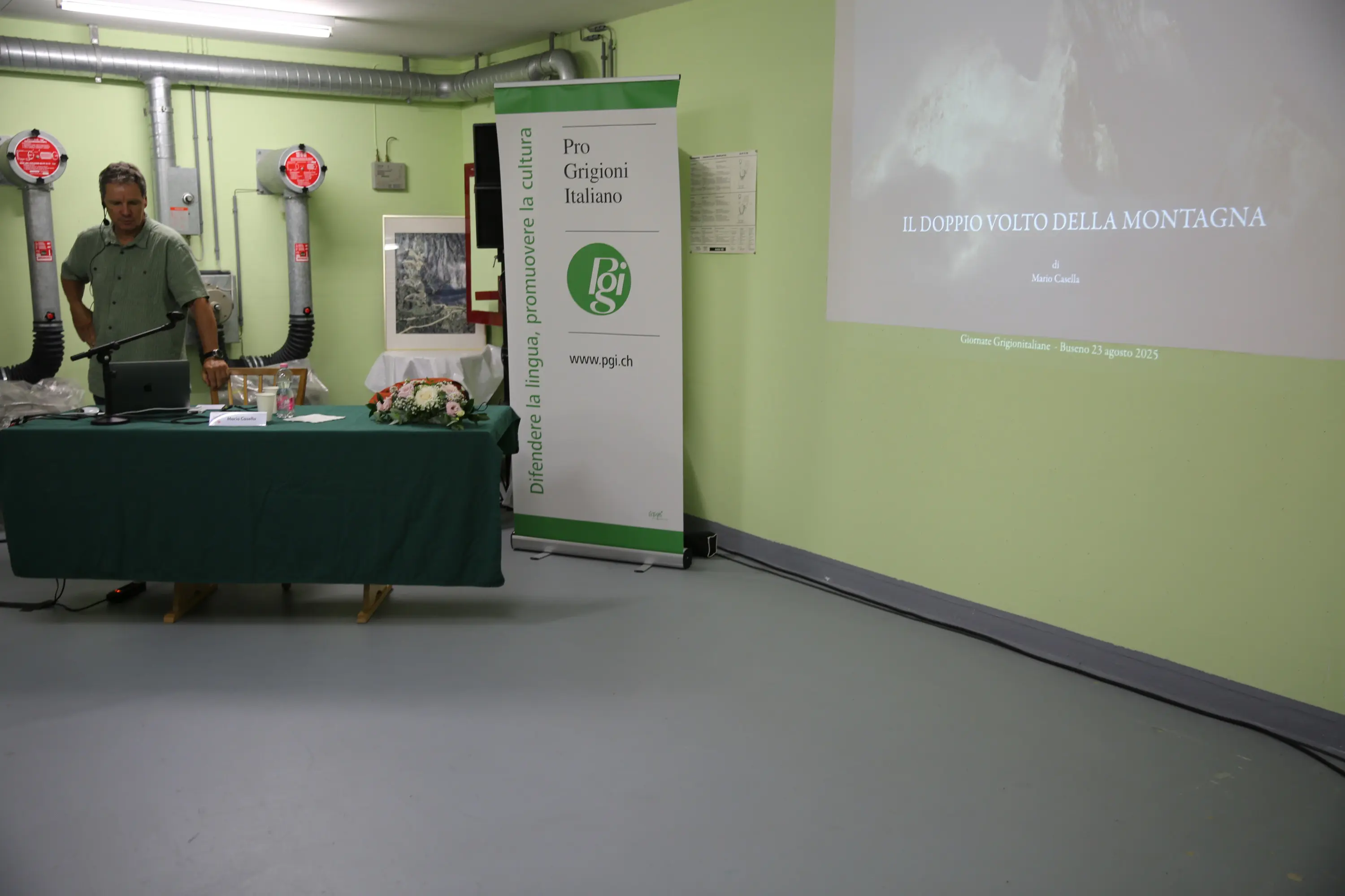 Un uomo si trova di fronte a un computer portatile su un tavolo verde, mentre presenta il tema "Il doppio volto della montagna" su uno schermo. Sullo sfondo si vede un banner con il logo dell'associazione Pro Grigioni Italiano. L'ambiente è semplice, con pareti verdi e apparecchiature visibili.