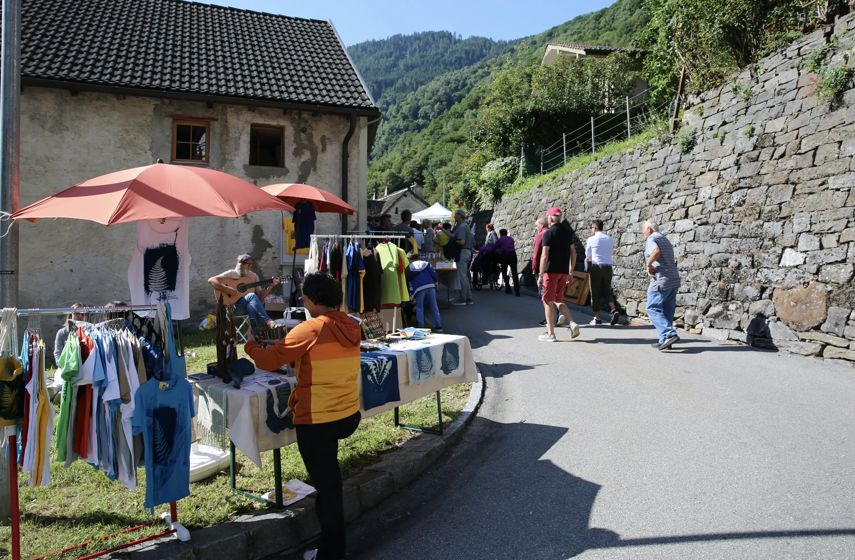 Mercatino all'aperto con bancarelle di abbigliamento colorato, ombrelloni rossi e persone che passeggiano lungo una strada di campagna. Sullo sfondo, case e colline verdi.
