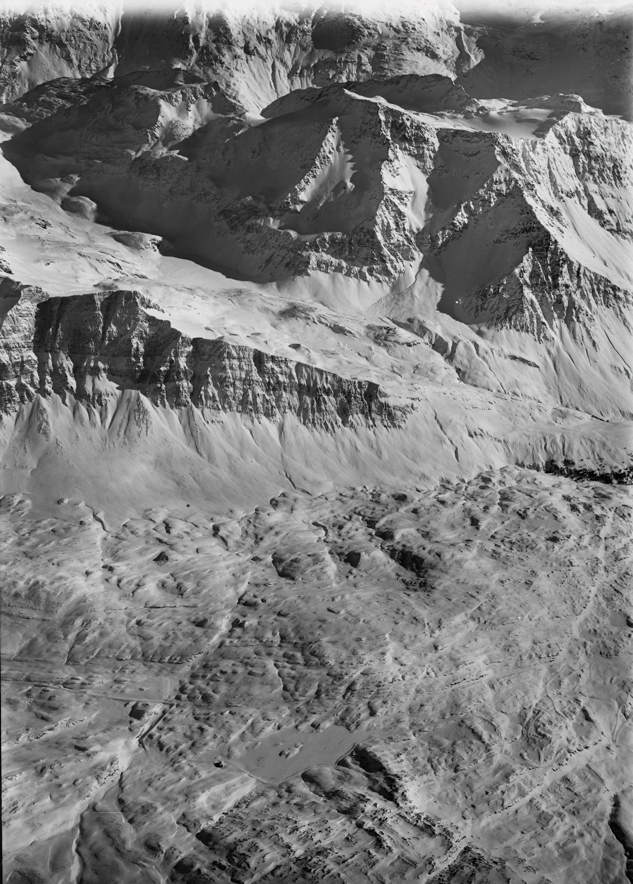 Vista aerea di un paesaggio montano imponente, caratterizzato da vette frastagliate e ripide scogliere. La scena è in bianco e nero, evidenziando le ombre e le texture delle rocce. La superficie sottostante mostra una variazione di forme geologiche.