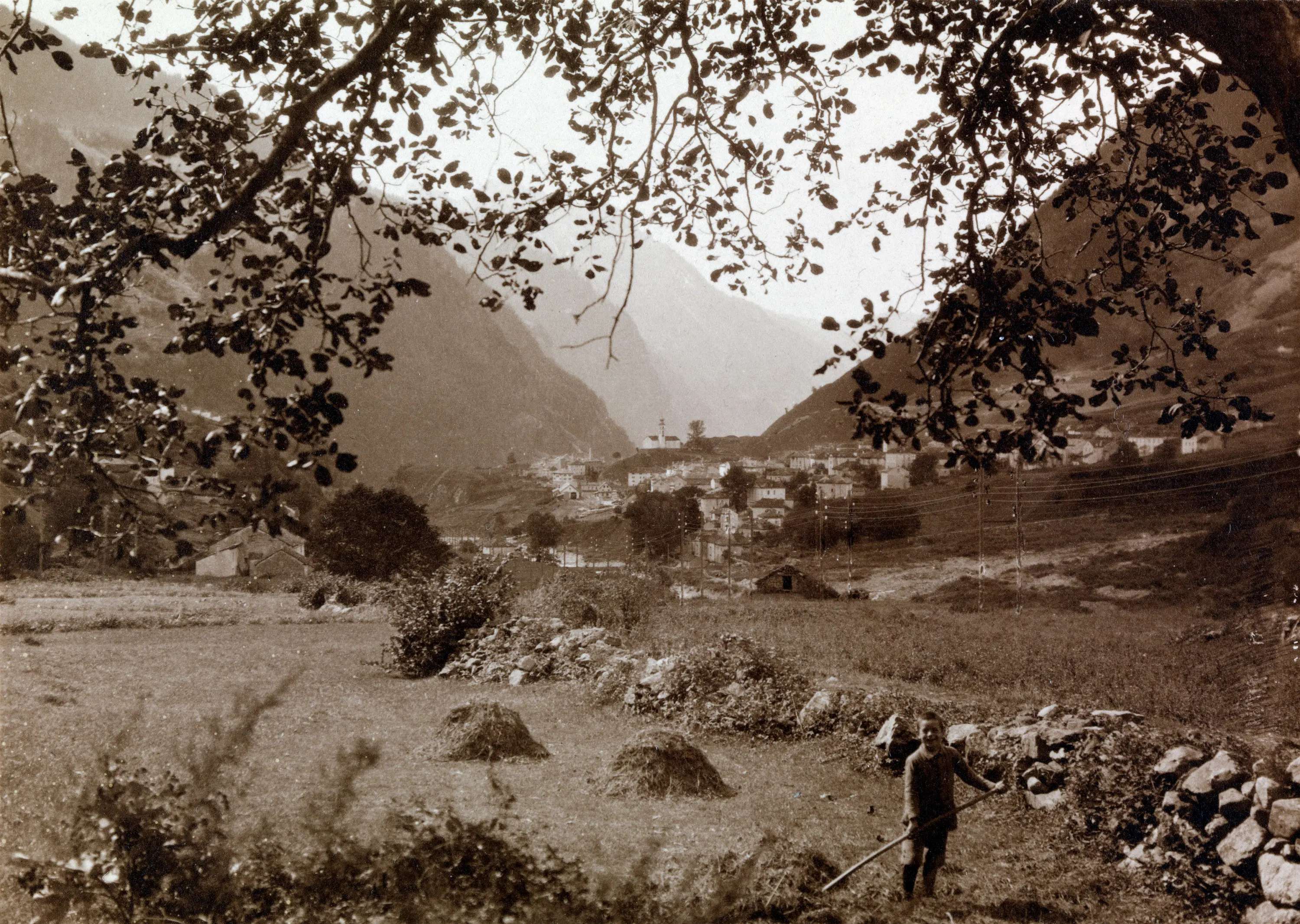 Paesaggio montano in bianco e nero con un contadino al lavoro in un campo, mentre sullo sfondo si intravede un villaggio circondato da montagne.