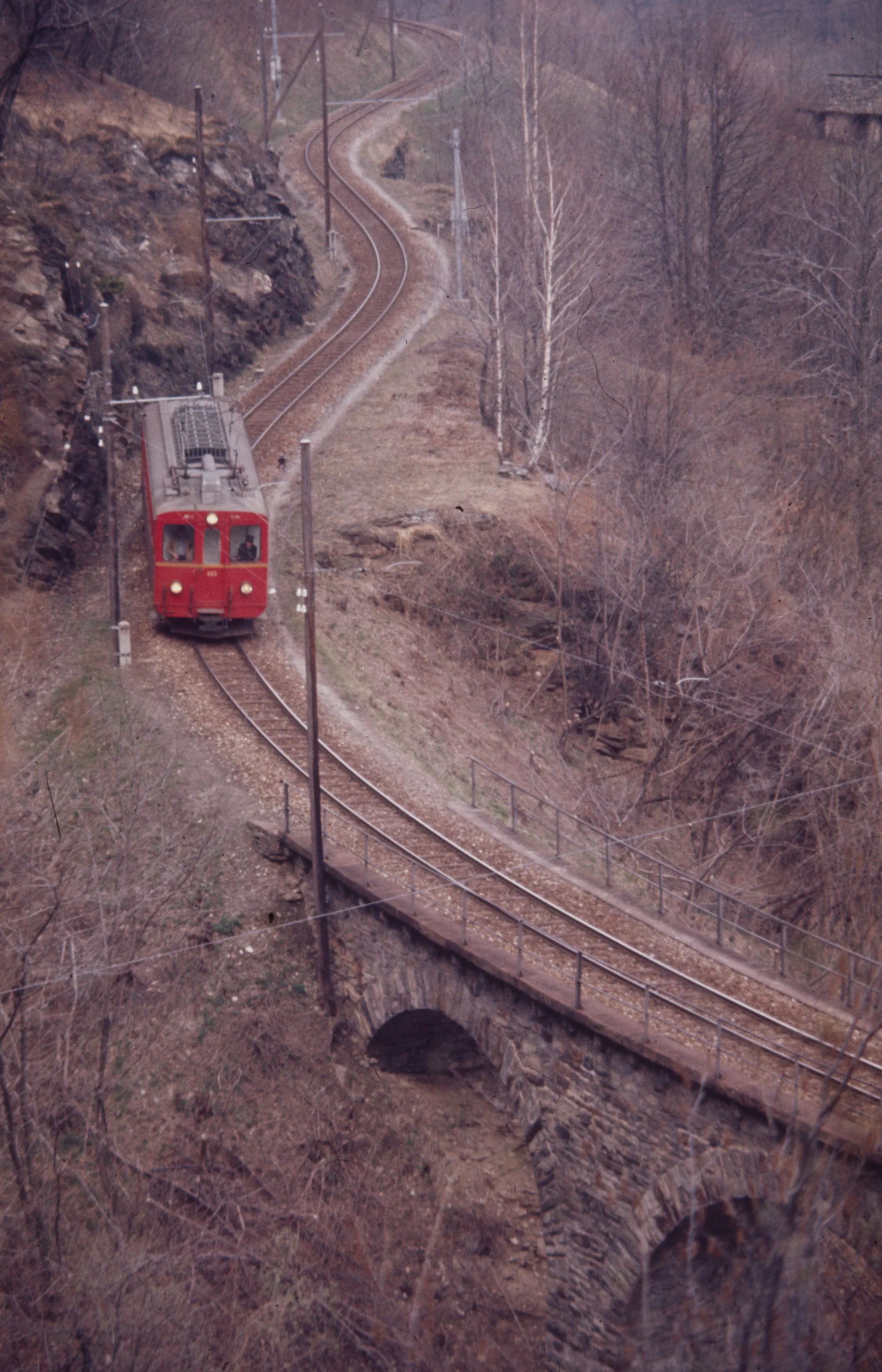 Un treno rosso percorre una ferrovia che si snoda tra la vegetazione secca, attraversando un ponte in pietra. L'ambiente circostante è collinoso e l'atmosfera è autunnale.
