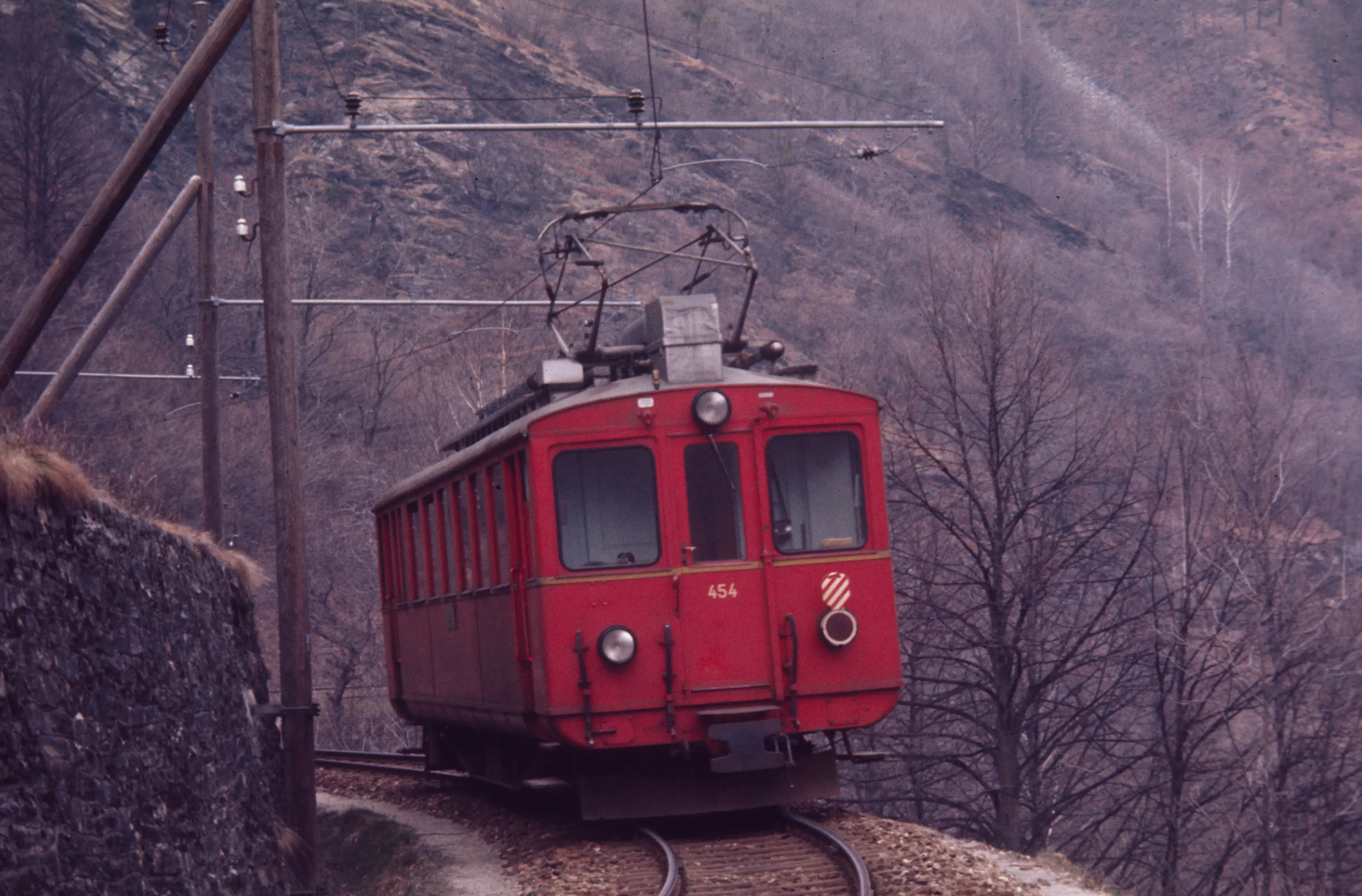 Un treno rosso percorre un binario tortuoso immerso in un paesaggio montano. Gli alberi spogli e le rocce circostanti suggeriscono un ambiente invernale.