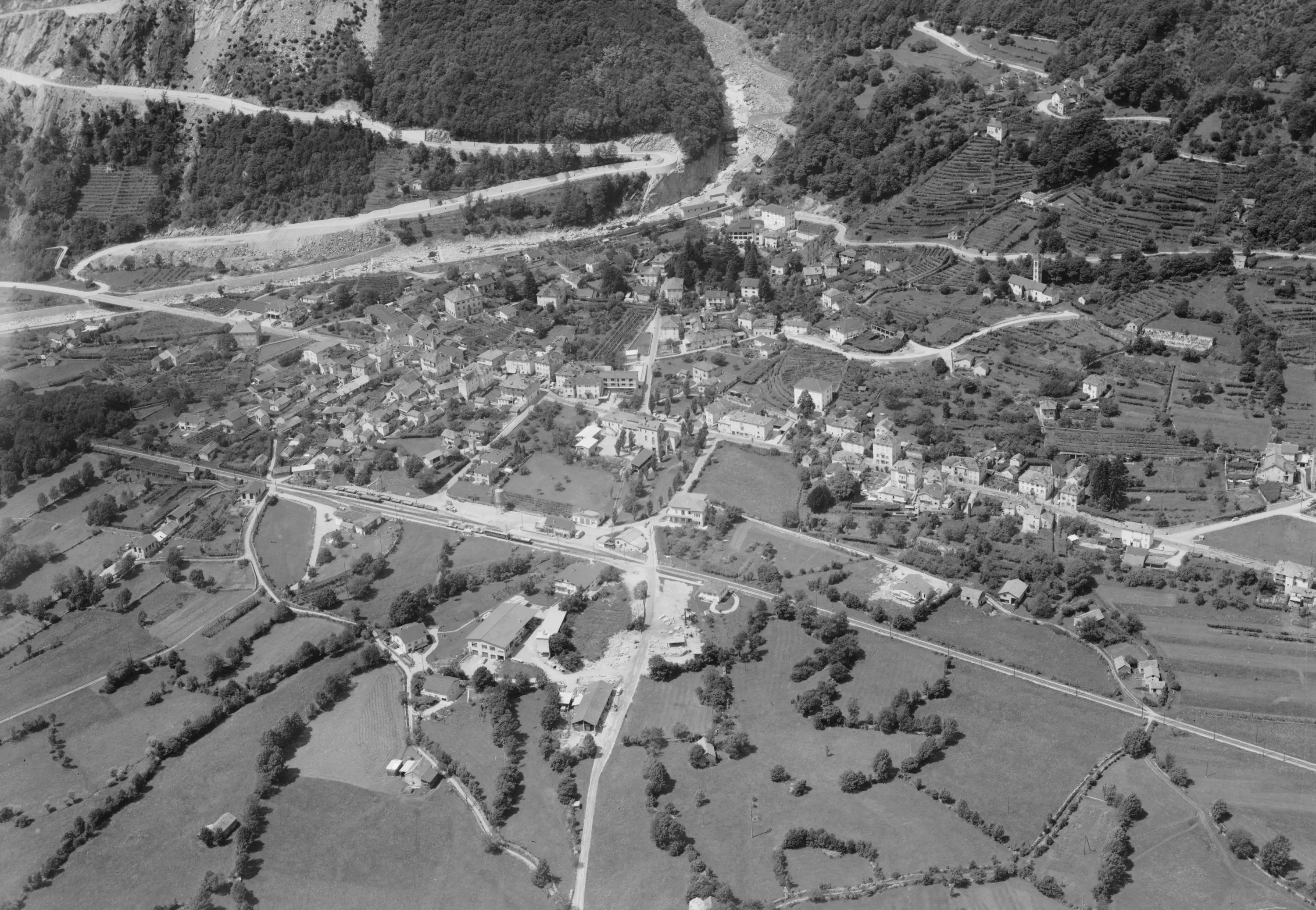 Veduta aerea in bianco e nero di un villaggio circondato da alberi e terreni agricoli, con strade che si intrecciano e case distribuite in tutto il panorama. Montagne verdi si ergono sullo sfondo.