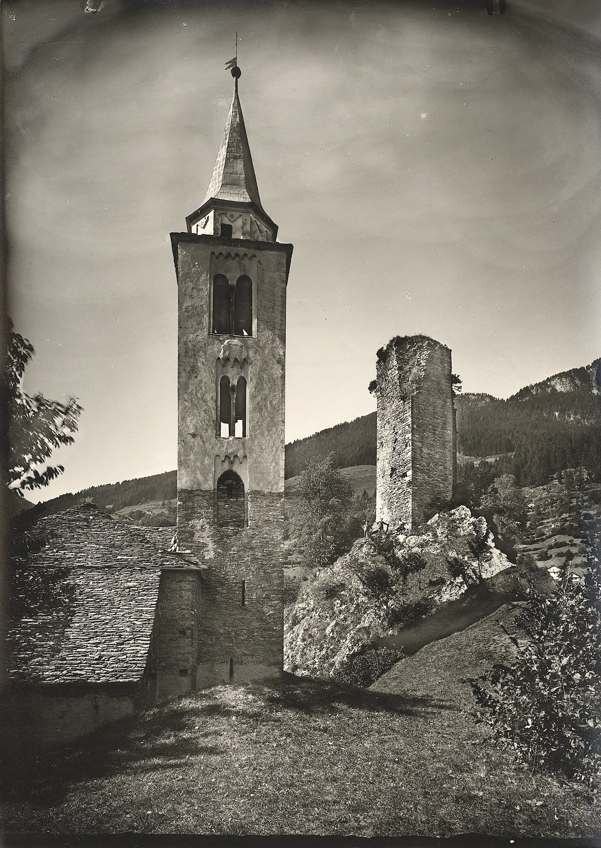 Un'antica torre campanaria con un tetto a punta si erge accanto a una rocca in rovina, circondata da una lussureggiante vegetazione. Sullo sfondo, si intravedono dolci colline e alberi.