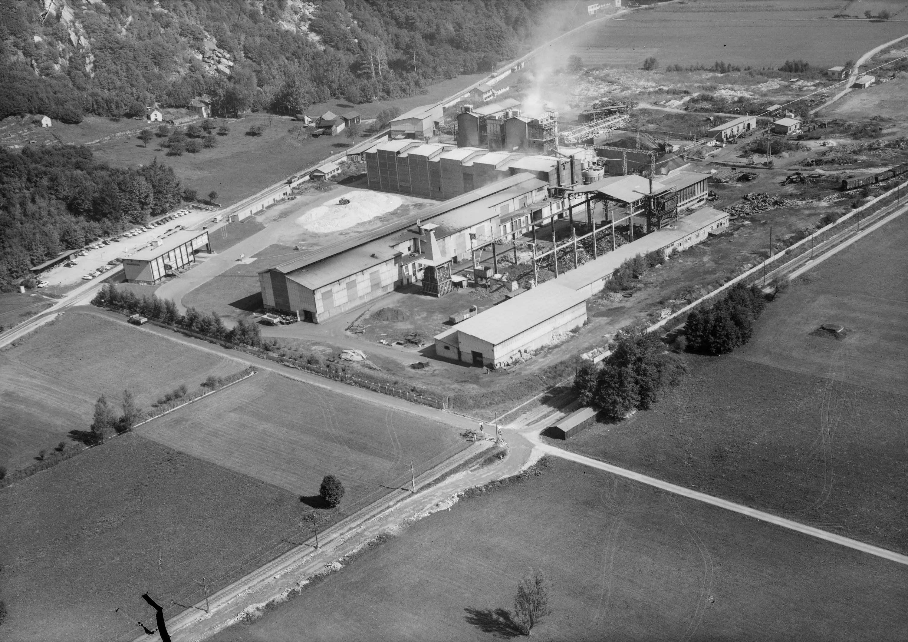 Immagine in bianco e nero di un impianto industriale circondato da campi verdi e boschi. Diverse strutture metalliche e silos sono visibili, con fumi che fuoriescono. Un'auto e una strada si trovano ai margini del sito.