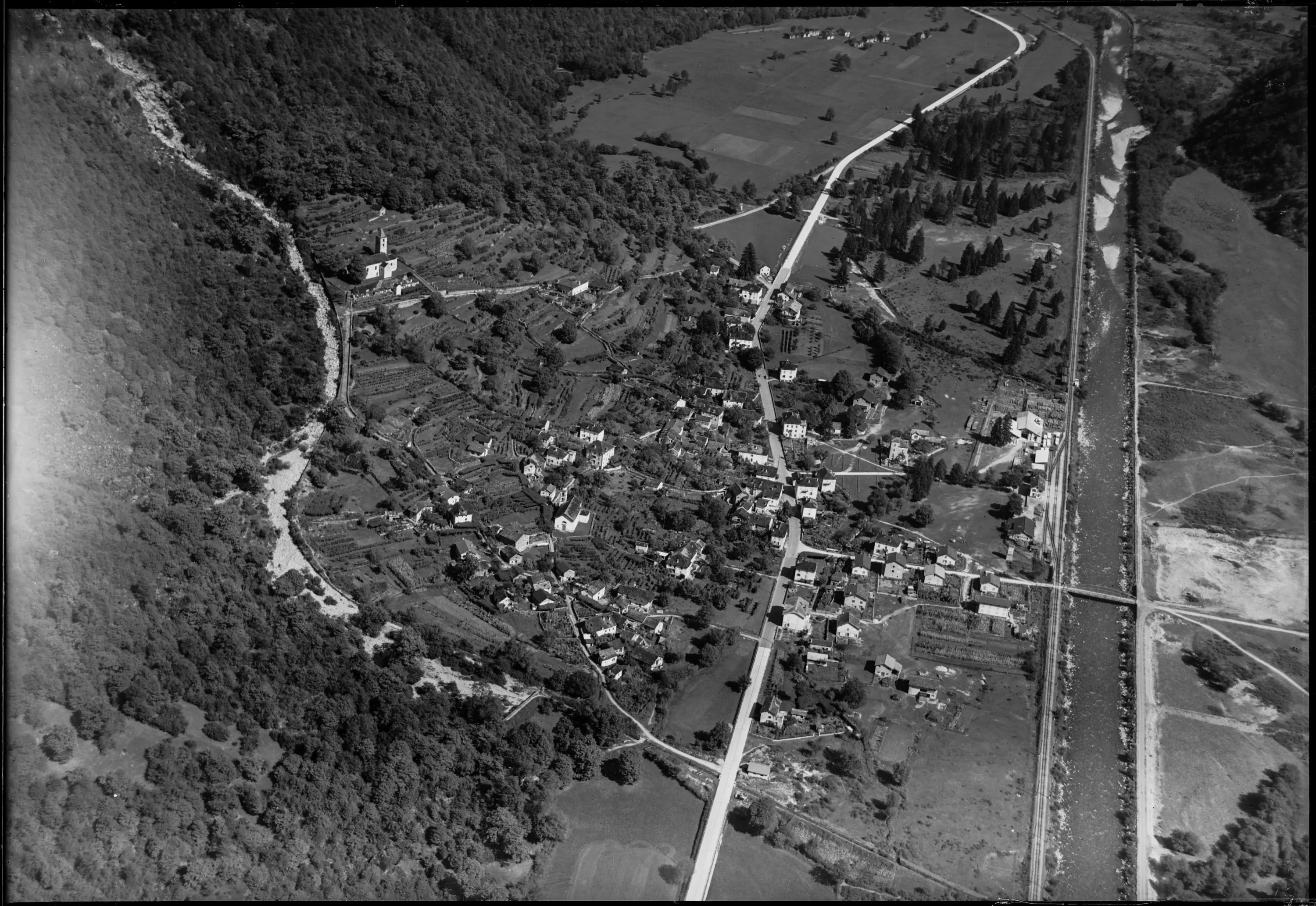 Veduta aerea di un piccolo paese circondato da colline e alberi, con strade che si snodano tra le case. Un fiume scorre lungo il perimetro del villaggio, mentre campi verdi si estendono nelle vicinanze.