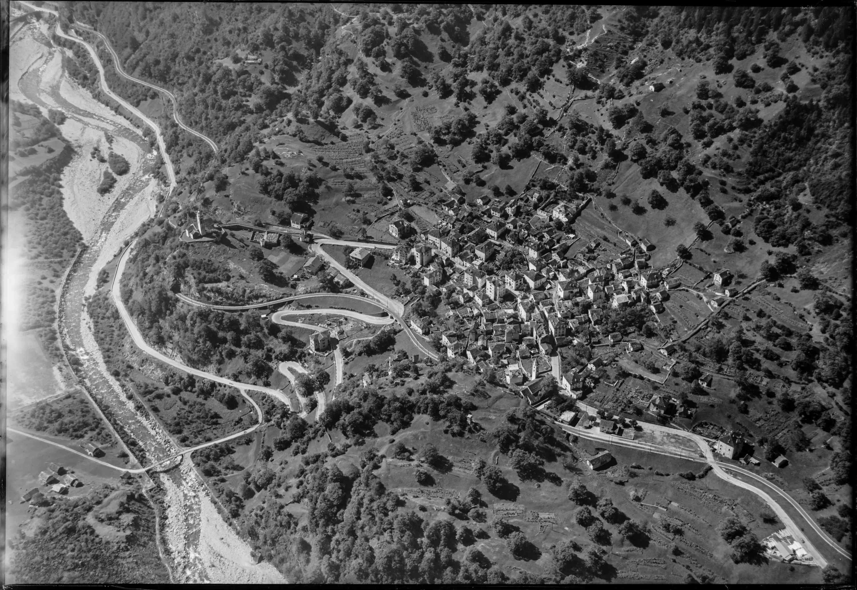 Veduta aerea di un piccolo paese circondato da alberi e colline, con strade sinuose che si snodano intorno. Un fiume percorre il paesaggio, mentre le abitazioni sono distribuite in modo compatto nel centro.