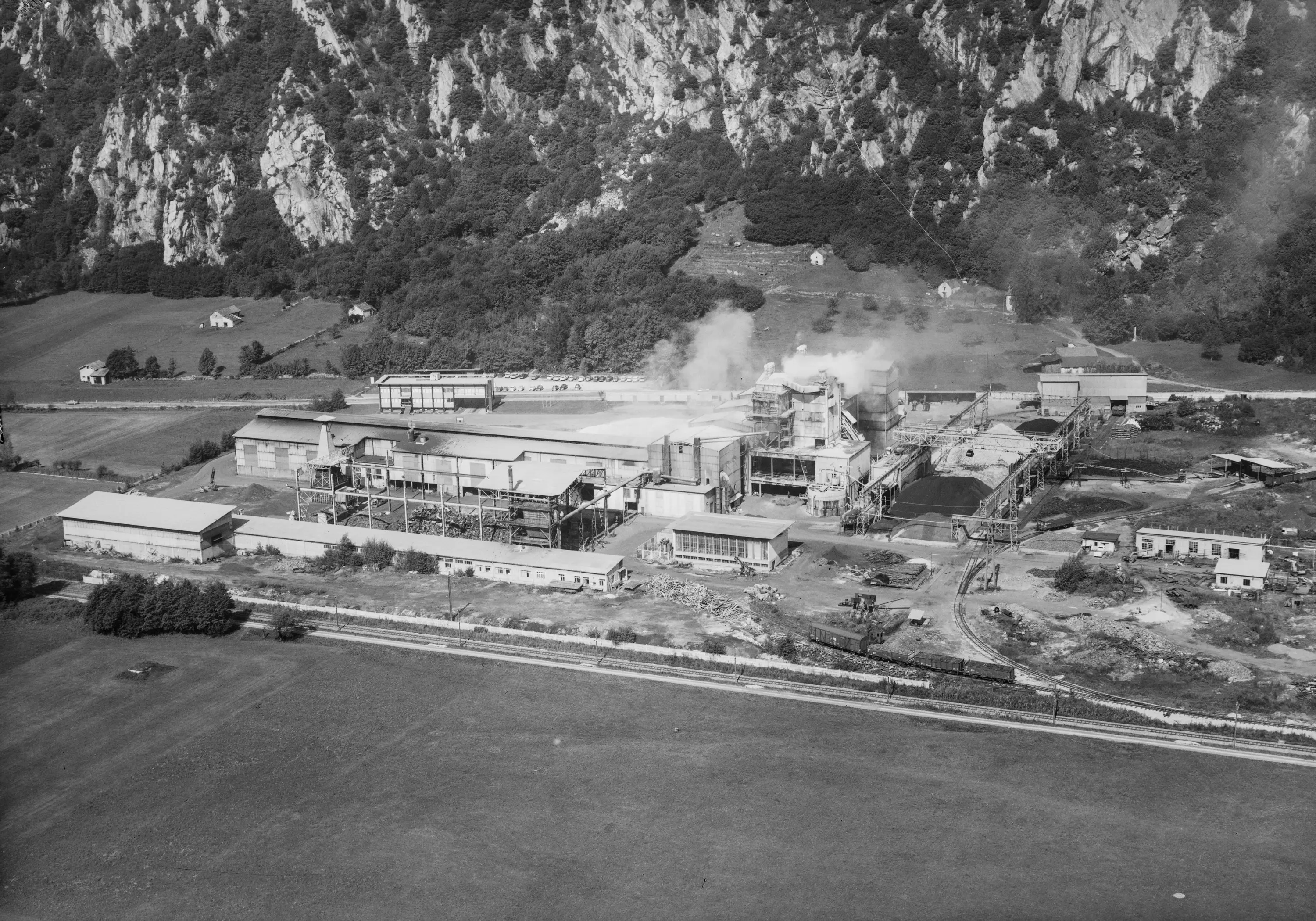 Un'industria vista dall'alto, circondata da montagne e vegetazione. Fumi bianchi escono da un camino. L'area circostante è caratterizzata da terreni verdi e alcune costruzioni.