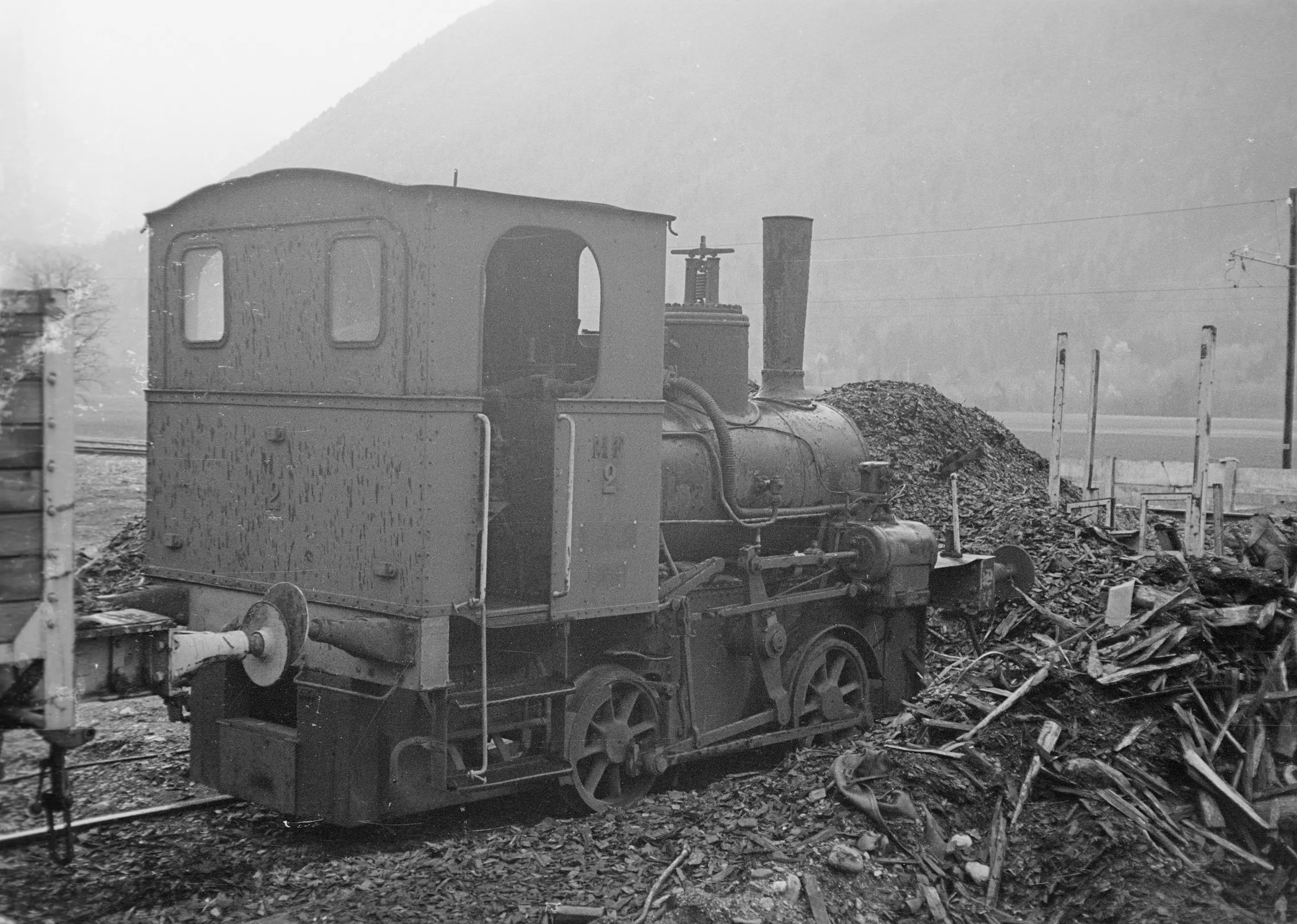 Locomotiva a vapore vintage parcheggiata vicino a un cumulo di legno, con una montagna sullo sfondo. Da notare il fumo che esce dal camino.