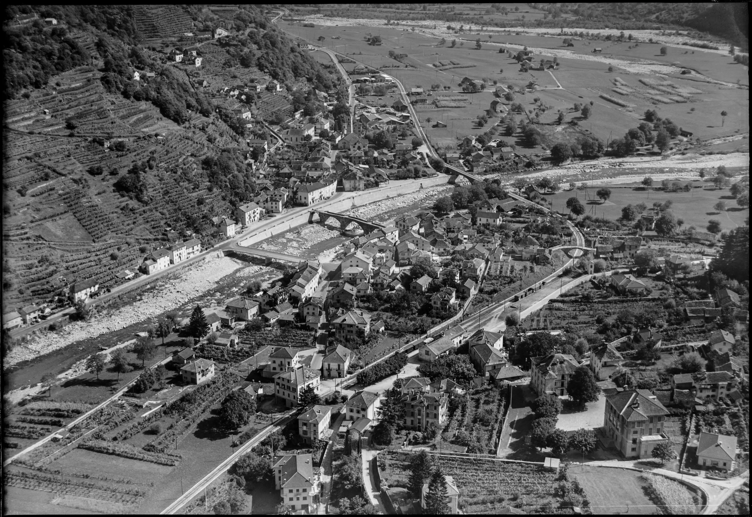 Veduta aerea di un villaggio circondato da colline, con case ben disposte lungo un fiume e strade che si intrecciano. Campi e vigneti si estendono attorno al centro abitato.