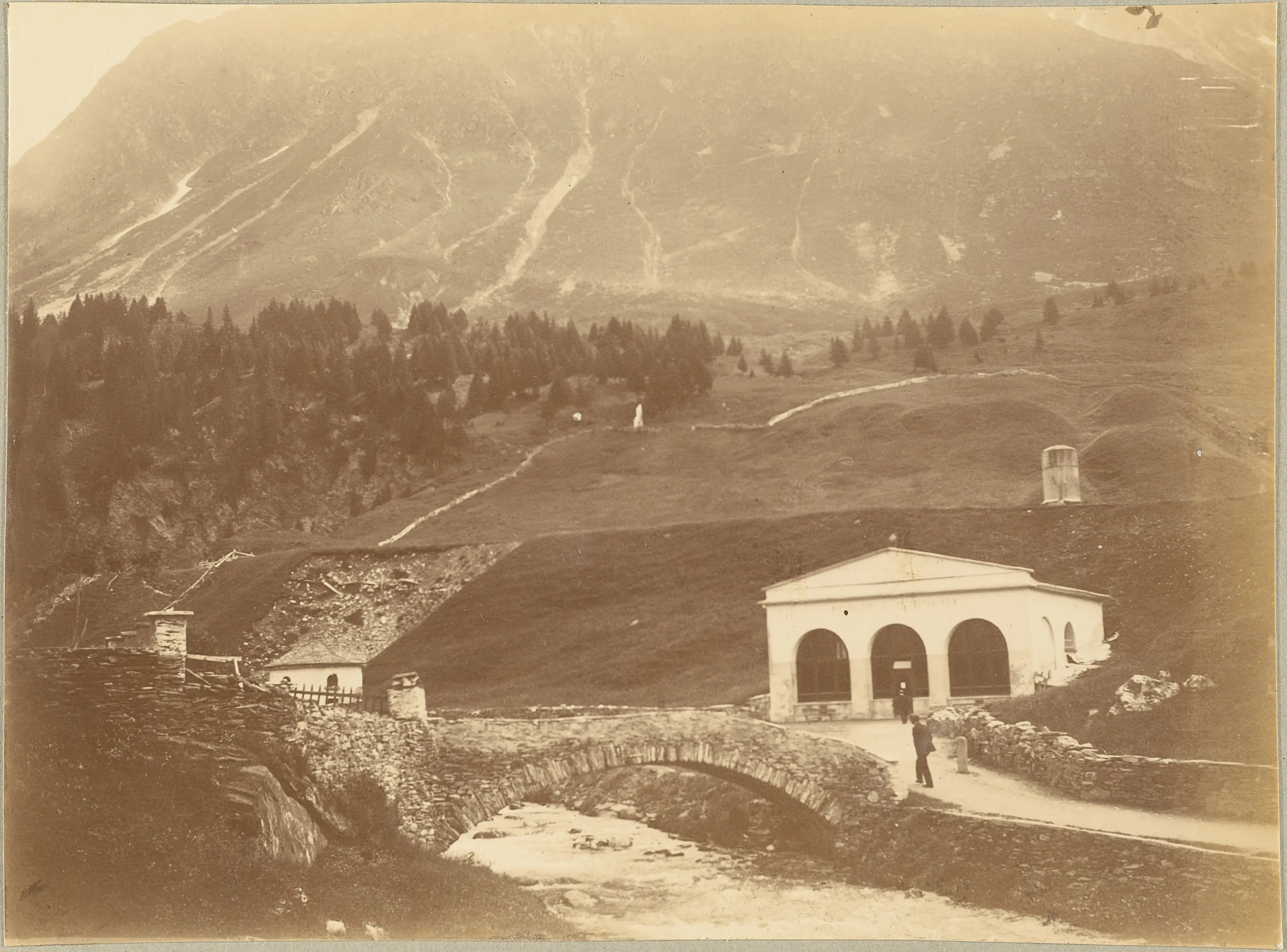 Un paesaggio montano con una struttura bianca in primo piano, una strada acciottolata, un ponte in pietra e alberi verdi. Sullo sfondo, si ergevano montagne imponenti e un cielo chiaro.