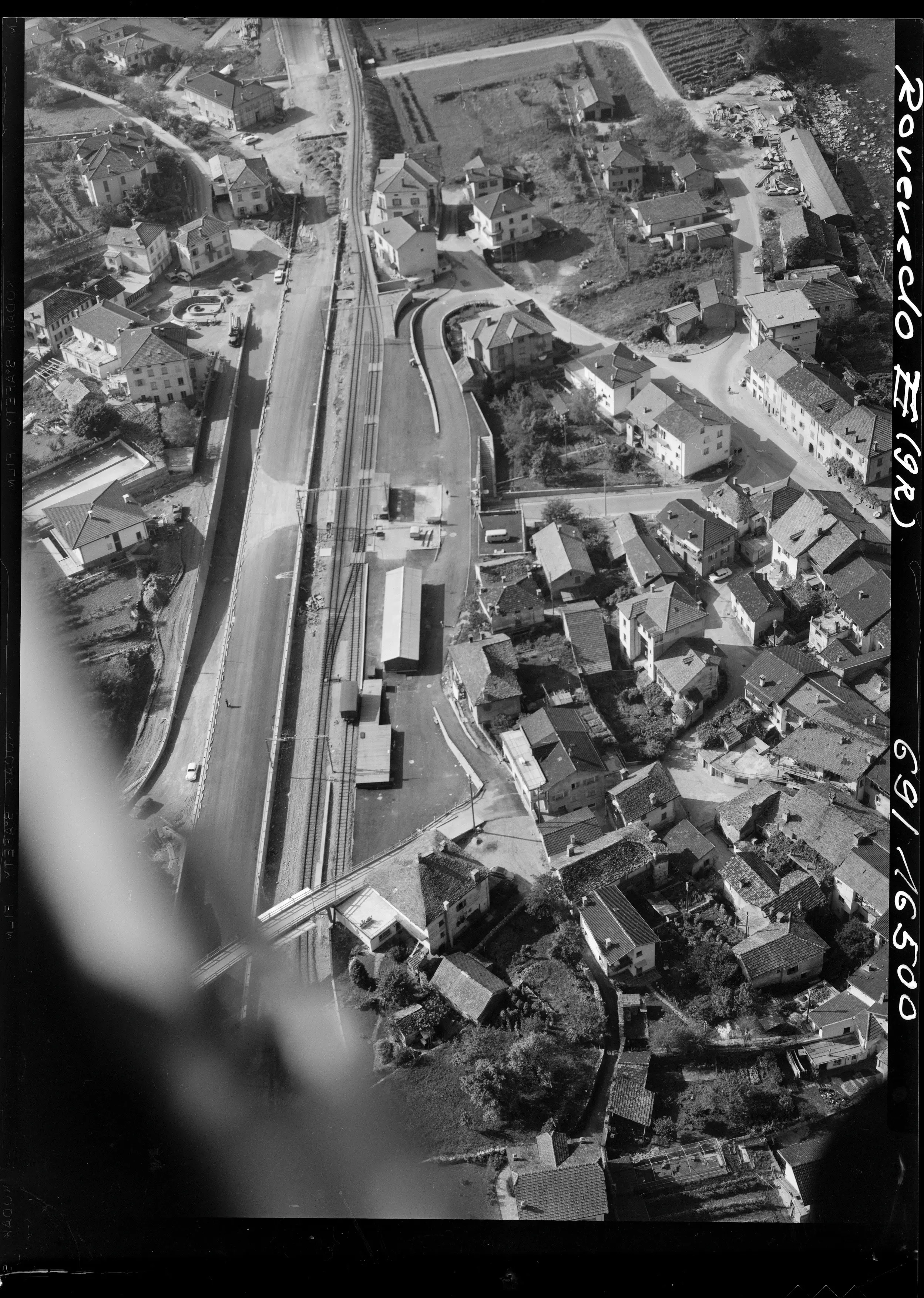 Vista aerea di Roveredo in Grigioni, con un tracciato ferroviario e piccole abitazioni circostanti. La zona mostra strade, edifici e un paesaggio montano in lontananza.