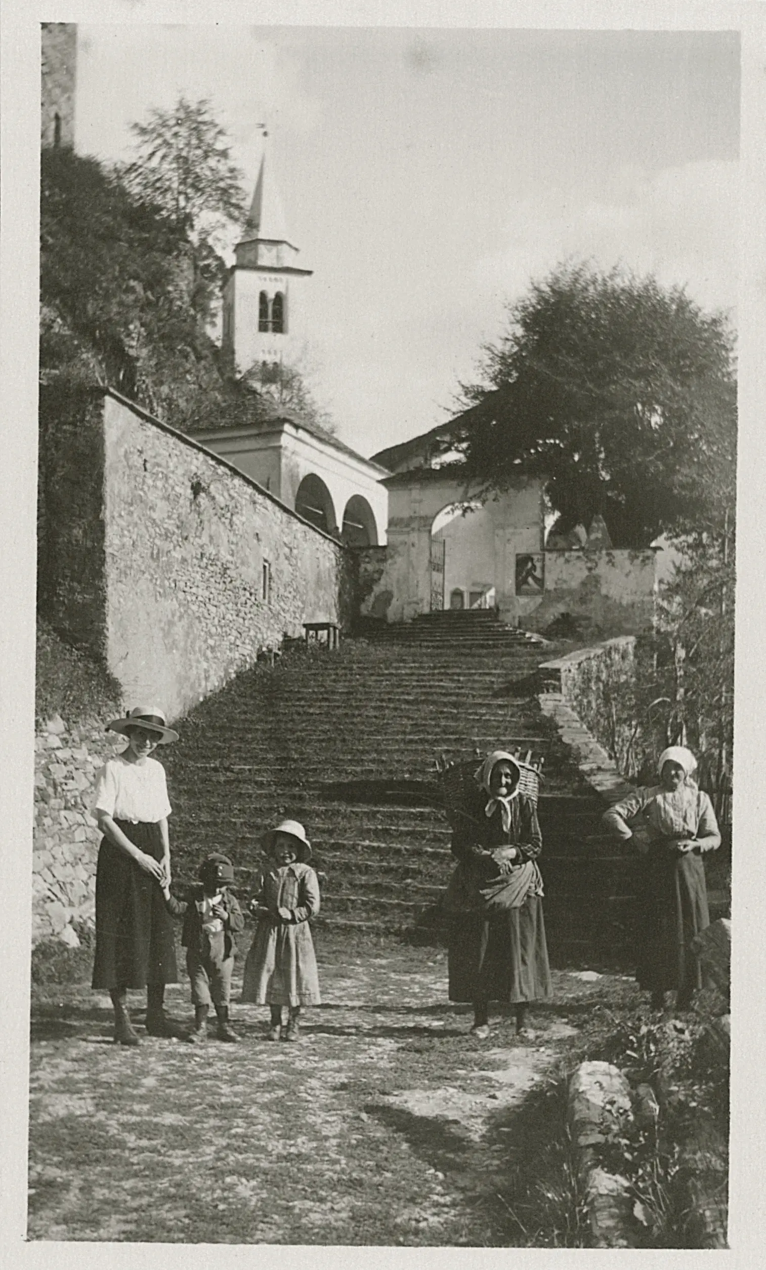 Due donne e due bambini in abiti tradizionali camminano su una scala di pietra. Sullo sfondo, si intravede una chiesa e edifici storici circondati da alberi. Il cielo è sereno e la scena evoca un'atmosfera di vita rurale.