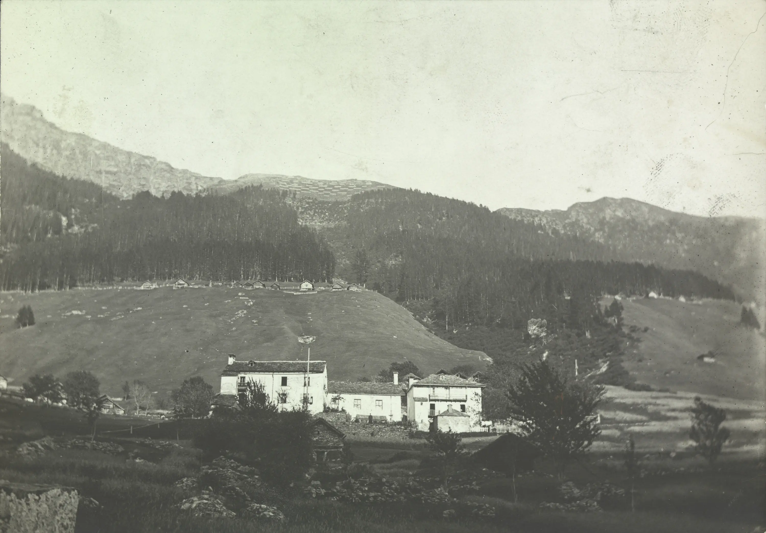 Un paesaggio montano in bianco e nero con una casa giù nel vallone, circondata da alberi e prati verdi. Le montagne si ergono sullo sfondo.
